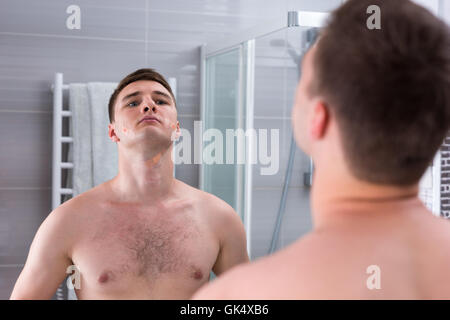 Giovane uomo cattivo dopo la rasatura con band-aids sul suo volto in piedi di fronte allo specchio nel moderno bagno rivestito di piastrelle a casa Foto Stock