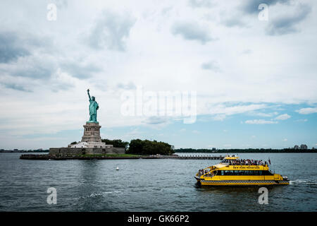 Una vista della statua della libertà nel porto di New York che mostra la statua con un giallo New York Water Taxi che passa in primo piano Foto Stock