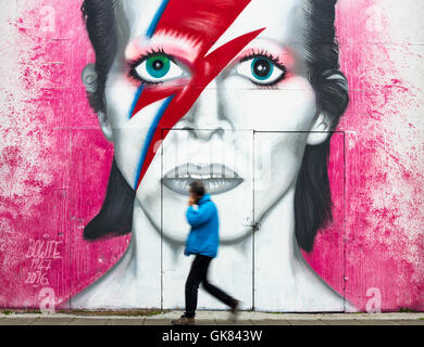 Newcastle upon Tyne, Inghilterra, Regno Unito. 19 Ago, 2016. Meteo: un tocco di colore su un livello di grigio il venerdì mattina in Gateshead come un uomo che parla al telefono cellulare passeggiate passato un grande murale di David Bowie vicino Il Sage Gateshead. Una banda di è prevista pioggia per spazzare in tutto il Regno Unito il venerdì. Credito: Alan Dawson News/Alamy Live News Foto Stock