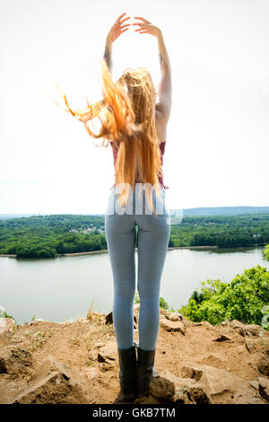 Vista posteriore della bella testa rossa in skinny jeans e parte superiore rossa, sulla cima della montagna con il vento nei capelli, braccia sollevate e un lago sottostante. Foto Stock