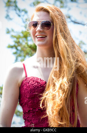 Bella testa rossa donna in occhiali da sole e parte superiore rossa, ridere mentre permanente sulla cima della montagna nella luce del sole dorato. Foto Stock