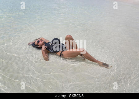 La donna in nero di un costume da bagno con spiaggia nera hat posa nelle acque di Osprey Cay, Exuma Cays, Isole Bahamas Foto Stock