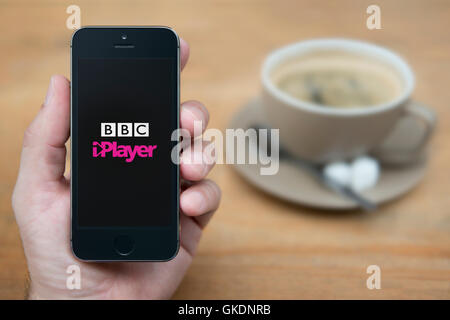 Un uomo guarda al suo iPhone che visualizza la BBC iPlayer logo, mentre sat con una tazza di caffè (solo uso editoriale). Foto Stock