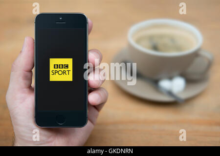 Un uomo guarda al suo iPhone che visualizza la BBC Sport logo, mentre sat con una tazza di caffè (solo uso editoriale). Foto Stock