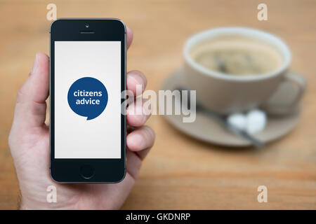 Un uomo guarda al suo iPhone che visualizza il Citizens Advice logo, mentre sat con una tazza di caffè (solo uso editoriale). Foto Stock