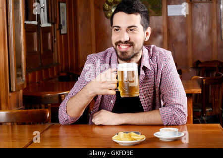 Ritratto di giovane uomo latino a bere birra e mangiare spuntini in un bar. In ambienti chiusi. Foto Stock