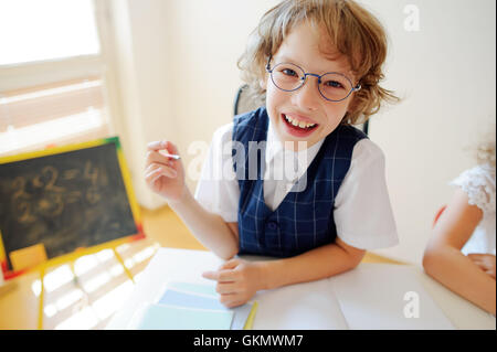 Funny piccolo scolaro in bicchieri si siede su un banco di scuola. Si tratta di un discepolo di una scuola elementare. In una mano al ragazzo di penna a sfera. Su Foto Stock