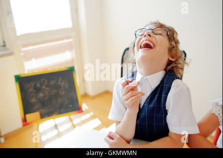 Funny piccolo scolaro in bicchieri si siede su un banco di scuola. Si tratta di un discepolo di una scuola elementare. In una mano al ragazzo di penna a sfera. Su Foto Stock