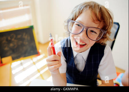 Funny piccolo scolaro in bicchieri si siede su un banco di scuola. Si tratta di un discepolo di una scuola elementare. In una mano al ragazzo di penna a sfera. Su Foto Stock