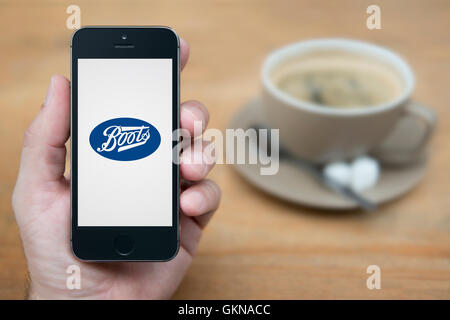 Un uomo guarda al suo iPhone che visualizza il logo di stivali, mentre sat con una tazza di caffè (solo uso editoriale). Foto Stock