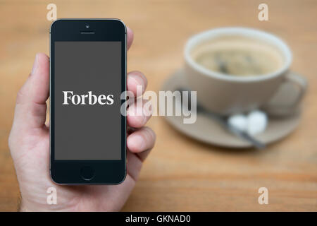 Un uomo guarda al suo iPhone che visualizza il logo di Forbes, mentre sat con una tazza di caffè (solo uso editoriale). Foto Stock