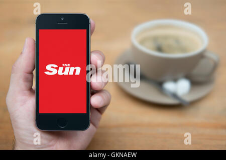 Un uomo guarda al suo iPhone che visualizza il logo Sun, mentre sat con una tazza di caffè (solo uso editoriale). Foto Stock