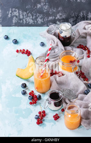 Varietà di melone e mirtilli frullati in vasetti di vetro e una bottiglia con yogurt e ribes rosso berry, servita sulla luce textu blu Foto Stock