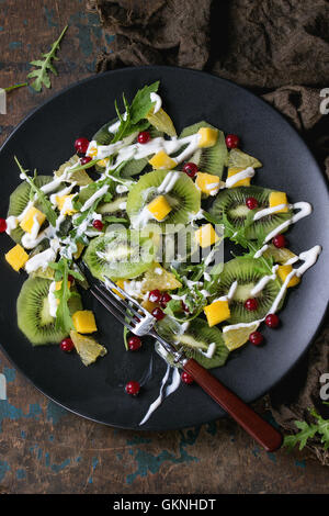 Macedonia di frutta con fette di kiwi, mango, arancio, rosso Ribes, rucola e dressing di yogurt, sulla piastra nera con la forcella e il cilicio rag Foto Stock