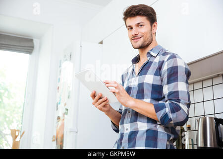 Sorridente Giovane Uomo in camicia a scacchi utilizzando compressa sulla cucina Foto Stock