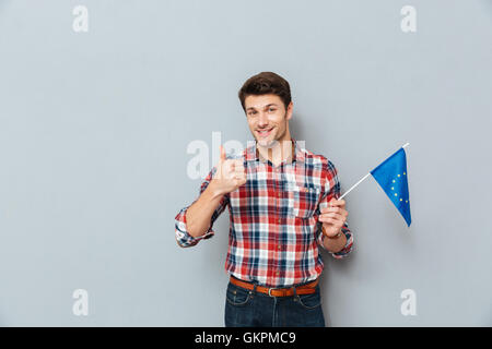 Felice giovane azienda bandiera della Unione Europea e mostra Thumbs up su sfondo grigio Foto Stock