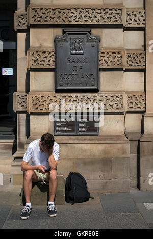 Un giovane uomo controlla il suo telefono mentre è seduto alla porta della Lloyds Banking Group Sede scozzese ex HBoS Foto Stock