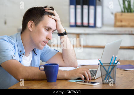 Ritratto di giovane uomo seduto al tavolo nella parte anteriore del computer portatile, assonnato, stanchi, affaticati o pigri per lavoro. Attraente business Foto Stock