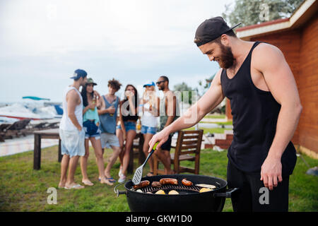 Bello barbuto giovane cottura sauseges alla griglia e verdura sul barbecue party Foto Stock