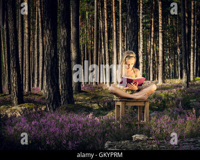 Bella ragazza bionda seduta su una sedia e la lettura di libro nella foresta. Foto Stock