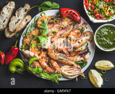 Piatto di gamberoni tigre arrostiti e pezzi di polipo con porri freschi, insalata, peperoni, limone, pane, pesto su sfondo nero Foto Stock