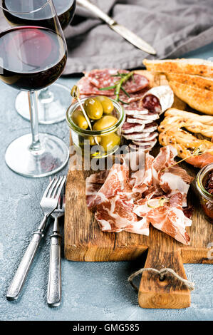 Antipasto a base di carne la selezione. Salame, prosciutto, grissini, baguette, olive e pomodori secchi, due bicchieri di vino rosso al di sopra di cemento grigio Sfondo testurizzata Foto Stock