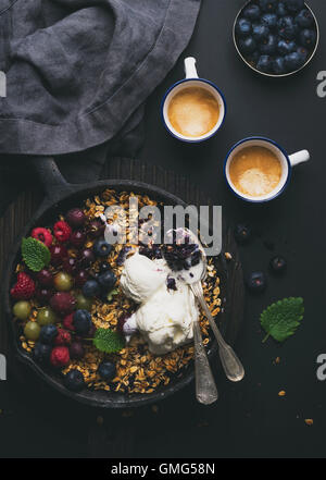 Una sana prima colazione. Oat granola sgretolarsi con frutti di bosco freschi, semi e gelati in padella di ferro padella in legno scuro, scheda di caffè su sfondo nero Foto Stock