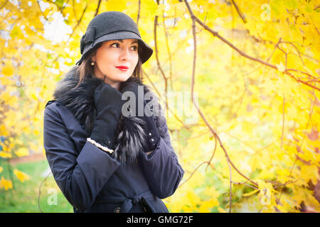 Ritratto di una giovane e bella donna in un mantello nero e hat su uno sfondo di colore giallo paesaggio autunnale Foto Stock