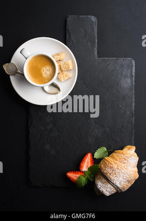 Tazza di caffè e croissant con fragole fresche in ardesia nera scheda di pietra su sfondo scuro Foto Stock