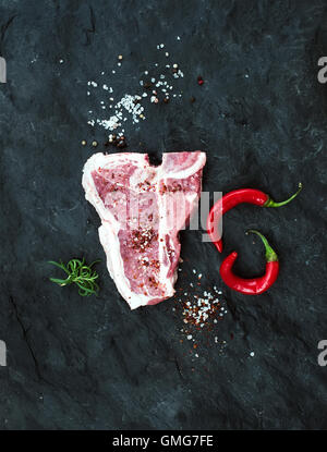 Materie carni fresche t-bone steak con spezie, peperoncino e rosmarino su ardesia nera sullo sfondo di pietra Foto Stock