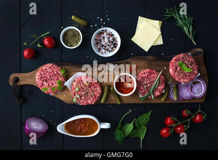 Ingredienti per la cottura di hamburger. Materie di macinato di manzo costolette di carne sul tagliere di legno, cipolla rossa, pomodori ciliegini, verdi, sottaceti, salsa di pomodoro, formaggio, erbe aromatiche e spezie su sfondo nero, vista dall'alto. Foto Stock