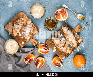 Tradizionale stile italiano home colazione. Latte in bicchieri, croissant alle mandorle su rustiche tavole di legno rosso e sanguinosa arance siciliane oltre il cemento tabella testurizzata Foto Stock