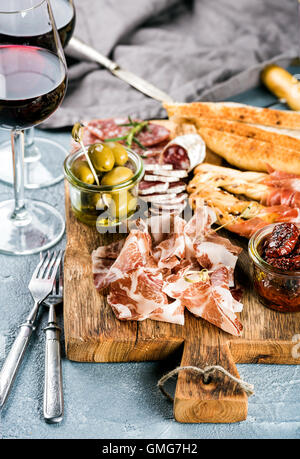 Antipasto a base di carne la selezione. Salame, prosciutto, grissini, baguette, olive e pomodori secchi, due bicchieri di vino rosso al di sopra di cemento grigio Sfondo testurizzata Foto Stock