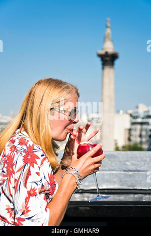 Ritratto verticale di una donna gustando un cocktail affacciato su Nelson's colonna in Trafalgar Square, Londra, sotto il sole. Foto Stock