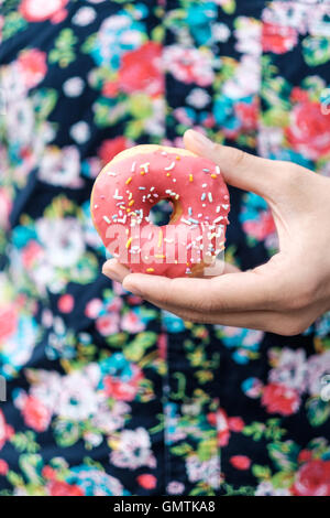 Mano che tiene una rosa cospargete ciambella contro un pulsante floreali up del modello camicia. Foto Stock