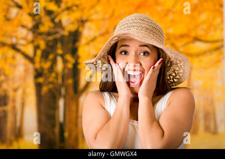 Giovane donna attraente indossando top bianco e di moda hat, toccando le guance con proprie mani urlando fuori, foresta autunno sfondo Foto Stock