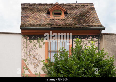 Carino piccola casa con ornamenti floreali su parete singola finestra e macchia verde nella parte anteriore. Foto Stock