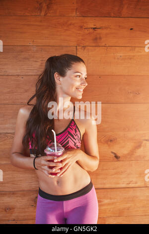 Colpo verticale di montare il modello femminile con un bicchiere di succo di frutta dopo la sessione di palestra, in piedi contro lo sfondo di legno. Sportive Foto Stock