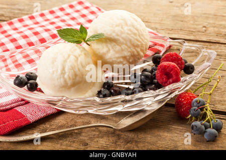 Due palline di gelato alla vaniglia con lamponi e mirtilli in recipiente di vetro sul tavolo rustico Foto Stock