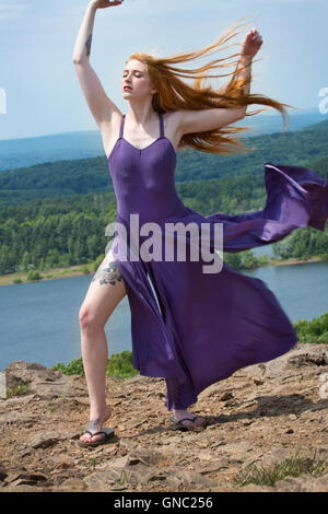Drammatico ritratto della bella testa rossa donna in fluenti vestito viola, sulla cima della montagna con il vento nei capelli e un lago sottostante. Foto Stock