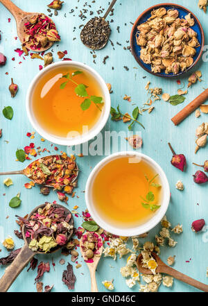 Due tazze di sano tè alle erbe con la menta, cannella, essiccato rose e fiori di camomilla in cucchiai su sfondo blu Foto Stock