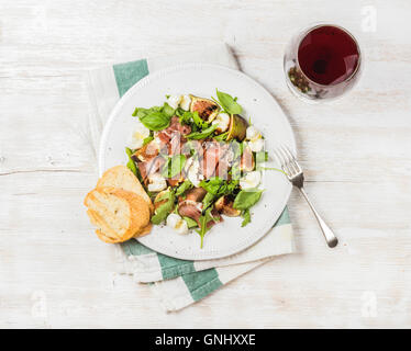 Il Prosciutto, rucola, basilico, fichi e insalata di mozzarella con un bicchiere di vino rosso su bianco sullo sfondo di legno, vista dall'alto Foto Stock