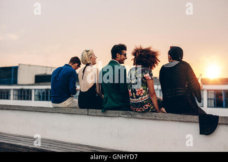 Vista posteriore del colpo di giovani uomini e donne seduti insieme sul tetto. Razza mista amici in relax sulla terrazza durante il tramonto. Foto Stock