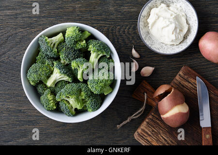 Una ciotola con broccoli freschi, i chiodi di garofano di aglio, patate e crema di formaggio su legno scuro dello sfondo. Brocoli gli ingredienti della zuppa. Foto Stock