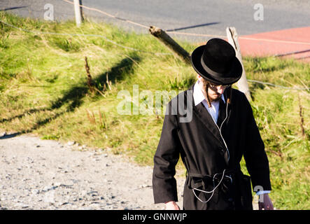 Un giovane ebreo Hasidic uomo con auricolari in, passeggiate in riva al mare in Borth, Wales, Regno Unito Foto Stock