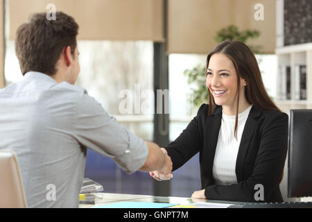 Felice imprenditrice di handshaking client con chiusura trattativa in un ufficio interno con una finestra in background Foto Stock