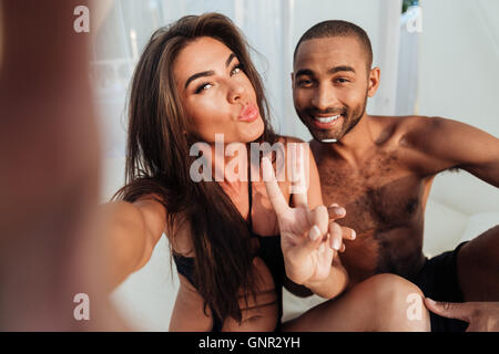 Giovane coppia sorridente in amore rendendo selfie seduti al lettino in spiaggia e mostra v segno Foto Stock