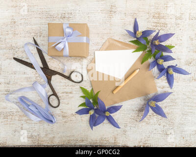 Confezione regalo, fiori blu, forbici, matita, nastro di raso e la busta con biglietto di auguri sul legno dipinto di bordo Foto Stock