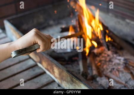 A partire il fuoco per la brace. Braccio con le tenaglie Long-Handled Impostazione Fire Closeup. Luminose fiamme e la combustione di legna. Foto Stock
