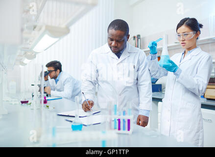 Femmina focalizzato scienziato asiatico blu di versamento di liquido nel pallone come maschio afro-americano di laboratorio di uno scienziato a scrivere i risultati Foto Stock